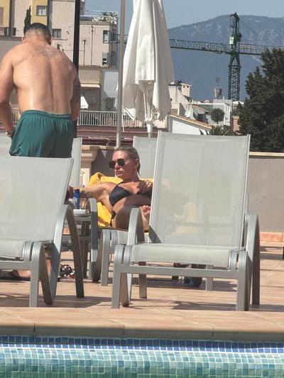The image captures a poolside scene at a building in Palma, Spain. In the foreground, a blue tiled swimming pool is visible. A woman is reclining on a sun lounger, wearing a black bikini and sunglasses. To her left, a blue bottle and a clear glass are on a small table. Behind her, a man with a tattoo on his back stands wearing green shorts. In the background, buildings and a construction crane are visible against a mountainous landscape under a clear sky.
