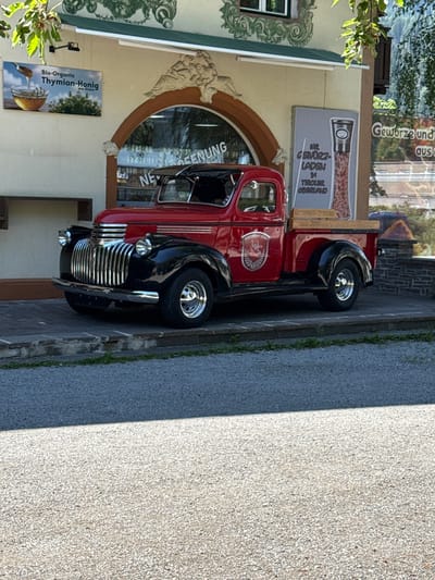 A vintage red and black Chevrolet pickup truck is parked on a paved walkway in front of a building. The truck has a chrome grille and shiny rims. A sign on the building behind the truck advertises "Bio-Organic Thymian-Honig" (Thyme Honey). Another sign displays a jar of spices and the text "Gewürz-Laden Tiroler Oberland". The building appears to be a shop, with a large arched window. Trees with green leaves are visible in the background, and part of a mountainous landscape is visible to the right of the building. The foreground shows a gravel road and a paved walkway with stone border. The overall scene suggests a quaint European village setting in Oetzerau, Austria.