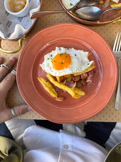 The image displays an overhead view of a prepared meal at Casa Vasco, situated in Aldoar, Foz do Douro e Nevogilde, Portugal. A round, salmon-pink ceramic plate occupies the central foreground, presenting a dish composed of a sunny-side-up fried egg, garnished with chopped chives, layered over golden-brown french fries, sliced sausages, and pieces of cured ham.
To the left of the plate, a person's left hand is visible, adorned with four silver rings: one on the pinky finger, one on the middle finger, and two on the ring finger. Above the hand, a small white ceramic cup containing a foamy coffee beverage rests within a brass-colored metal holder with a handle. This holder is placed on a disposable paper liner featuring the text "CASA VASCO."
On the right side of the main plate, a silver four-tine fork is positioned vertically. In the upper right background, a portion of a second copper-toned metal dish or pan is partially visible, containing remnants of food, including fries and a fried egg, alongside two silver spoons. The table surface is light-colored with a repeating pattern of small reddish-brown star-like designs.
The lower section of the image reveals parts of a person's attire: a white collared shirt with a horizontal navy blue stripe, and a section of a green or khaki-colored bag in the bottom-left corner.