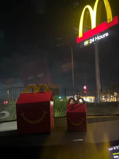 The image shows a nighttime scene at a McDonald's restaurant, likely in Johannesburg, South Africa, although specific location information is not visible. Two Happy Meal boxes are placed on a surface, possibly a car dashboard. In the background, a McDonald's sign with the golden arches and "24 Hours" text is illuminated. Outside the window, city lights are visible. The time 21:37 is shown on a device in the top right corner. The boxes are red with a golden smile on the front and golden handle.