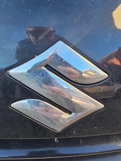 The image provides a close-up view of a chrome vehicle emblem, set against a dark, likely black, and visibly dusty and scratched metallic surface, characteristic of a car's exterior. The emblem is a stylized, angular design, strongly resembling the modern logo for Mahindra, featuring interconnected, diamond-like shapes that form a unique 'M' or 'W'.
The polished chrome surface of the emblem reflects its surroundings vividly. Prominently visible are reflections of a bright blue sky and what appears to be part of a building or another vehicle in the background. Most notably, the distinct reflection of the photographer's arm, hand, and possibly part of their body, holding what seems to be a camera or phone, can be seen across the emblem's surface.
The dark paintwork around the emblem shows a significant layer of dust, small specks of dirt, and several fine scratches, indicating that the vehicle has been exposed to the elements and has not been recently cleaned. The bright reflections suggest the photograph was taken during daytime, under clear or brightly overcast conditions.
The scene captures a static object on a vehicle, with no specific activity taking place other than the act of photography. The wear and tear on the vehicle's surface suggest it is an older model or has seen considerable use. The location context provided is Holywell, United Kingdom, implying this vehicle is situated there. There is no explicit text visible other than the symbolic form of the emblem itself.