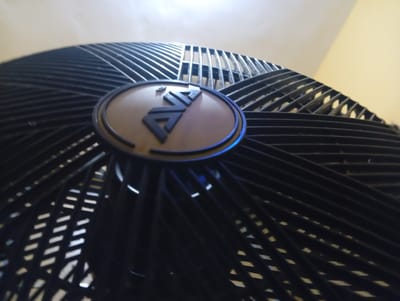 A close-up, low-angle shot shows the front grille and logo of a black oscillating fan. The grille is composed of thin, parallel black bars that fan out from the center, creating a radial pattern. The fan's logo is a circular emblem with a brown wood-grain texture at its center, featuring a stylized black "AVA" insignia. The "AVA" logo is outlined with a thin, bright blue light. The background consists of a lightly textured, pale yellow wall and a lighter, off-white ceiling, suggesting an indoor environment. No people are visible in the image. The scene is static and the fan appears to be off. The image was taken in Ukukwa, Nigeria.