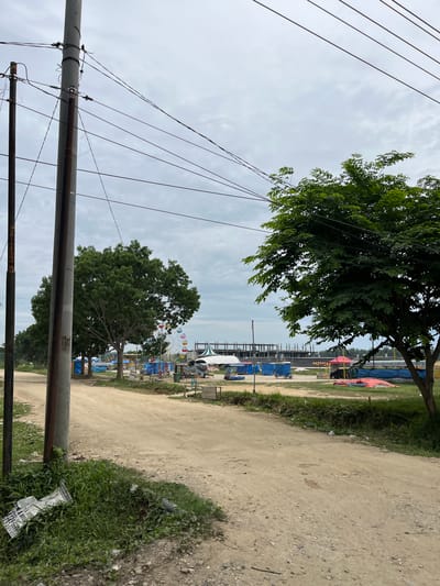 This image captures a street scene in Bundar, Indonesia, under an overcast sky. A dirt road dominates the foreground, with a prominent utility pole on the left side of the frame adorned with numerous electrical wires that crisscross the sky. To the right of the road, a lush green tree with dense foliage extends into the frame.
In the middle ground, a Ferris wheel with colorful pods stands partially obscured by trees. Beyond the Ferris wheel, several temporary structures covered in blue tarps are visible, suggesting a market or a gathering. Some small carts or stalls are also present. In the background, the skeletal framework of a large building under construction, possibly a stadium or industrial facility, is discernible against the cloudy sky. The ground appears to be a mixture of dirt and sparse grass, with some scattered debris near the utility pole. The overall atmosphere suggests a developing area, possibly on the outskirts of a town or city.