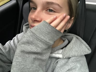 A young woman in a gray zip-up hoodie is shown from a close-up, tilted perspective. She partially obscures her face with her hand, revealing green eyes and a nose ring. Her nails are manicured with light pink polish. Parts of a car interior are visible in the background, suggesting she is sitting in a car seat. A silver chain hangs from her neck.