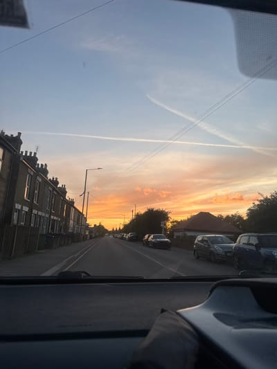 The image shows a street scene in the Borough of Swale, United Kingdom, during sunset. The view is from inside a vehicle, looking forward through the windshield. The sky is a mix of blue and orange hues, with wispy clouds and contrails visible. On the left side of the street, there is a row of brick terraced houses with chimneys and windows. Some green and blue bins are lined up along the pavement in front of the houses. On the right side of the street, there are more houses and trees, and several cars are parked along the roadside. The street itself is paved and appears to be a residential road. Streetlights are visible along the road. The overall impression is a quiet suburban street at the end of the day.