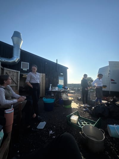 The image depicts an outdoor, commercial or event-related scene, stated to be in Northallerton, United Kingdom, under a clear blue sky with the sun low on the horizon. Four individuals are present, all appearing female.
On the left, a dark-colored, single-story building with horizontal siding is visible. A large, L-shaped corrugated metal exhaust duct extends from its left side, above a rectangular metal vent. A wooden door is open on the building's left facade, and another door is visible further right.
In the left foreground, one individual is seated on a wooden outdoor chair, facing right. An orange-pink insulated cup is on a surface next to them. Standing to her right, a second individual in a light-colored shirt and dark trousers stands facing left-front.
Central to the image, a makeshift table is positioned near the building, holding items including spray bottles and blue containers. A blue bucket and a black bucket are on the gravel ground near this table.
To the right, a large white van is partially visible. Text on its side includes "pple food" and "Hyd & All," with a license plate ending in "YNZ". Two additional individuals stand near the van. One, dressed in dark attire, faces left. The other, wearing a white shirt and a dark apron, stands with hands on her hips, facing right-front.
The ground consists of gravel. Various items are scattered, including black refuse bags near the van, a green plastic crate on its side containing a white plastic container, a large metal pot, and smaller debris. The lighting indicates strong, low-angle sunlight creating pronounced shadows. The overall scene suggests a functional outdoor workspace, potentially related to food service or an event.