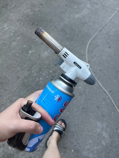 This image shows a close-up view of a person's hands holding a butane torch connected to a blue aerosol can. The torch is white and gray with "FLAME GUN" printed on it. The aerosol can has blue text and graphics, including a snowflake icon and some Cyrillic lettering that appears to be instructions or warnings. A person's feet, wearing sandals, are visible in the background, suggesting an outdoor setting. The ground is a gray, textured surface, possibly concrete or asphalt. A thin, transparent tube or wire is also visible running across the ground. The lighting suggests it is daytime, but the weather is not discernible. The overall scene appears casual and practical, with the torch and can suggesting a task involving heat or flame. The text on the can, which is partially legible in Russian, includes phrases like "SEASONAL," "under pressure," "out of reach of children," and warnings about heat and fire.