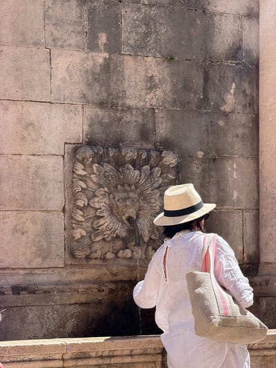 A person wearing a white linen shirt, a straw hat with a black band, and carrying a canvas tote bag with pink and blue stripes, stands before a stone wall with a carved lion's head fountain. Water flows from the lion's mouth into a basin below. The wall is constructed of large, rough-hewn stone blocks, showing signs of age and weathering. The setting is likely a public space within the historic city of Dubrovnik, Croatia, possibly near the Pile Gate or within the Old Town. The sunlight suggests it is daytime.