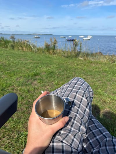 In the city of Am Salzhaff, Germany, a person sits in a chair outdoors, holding a metal cup of coffee and a cigarette. The person is wearing plaid pants. In the background, there is a body of water with several boats, and beyond that, a grassy field and a blue sky with some clouds. The scene suggests a peaceful moment of relaxation by the water.