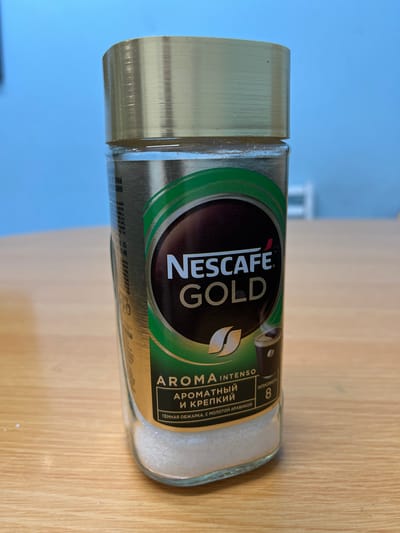 A clear glass jar containing white, granular coffee is centered in the frame, resting on a light-brown wooden surface. The jar has a metallic gold lid with horizontal ridges. A green and black label is affixed to the front of the jar, featuring the "NESCAFE GOLD" logo in white and red lettering. Below the logo, Russian text describes the coffee as "AROMA INTENSO", "АРOMATHЫЙ И КРЕПКИЙ" (aromatic and strong), and "ТЁМНАЯ ОБЖАРКА, С МОЛОТОЙ АРАБИКОЙ" (dark roast, with ground arabica). An intensity rating of "8" is also visible on the label. The background is a soft blue wall. The image appears to be taken indoors, possibly in a kitchen or on a desk, on a table. There are no people present in the image. The scene does not depict any specific location in Murmansk, Russia.