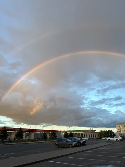 An expansive double rainbow arches across a dramatic, cloudy sky, dominating the left side of the frame. The sky features a mix of dark, heavy clouds and patches of brighter, lighter clouds, with hints of golden light breaking through, suggesting either sunrise or sunset and recent rain. The primary rainbow is vibrant and clear, while a fainter, secondary rainbow is visible outside of it.
On the right side of the image, there is a large asphalt parking lot with multiple cars parked in designated spaces. Beyond the parking lot, a row of buildings stretches horizontally. The most prominent building on the right has a long red sign with Cyrillic text that reads "АВТОМОЙКА САМООБСЛУЖИВАНИЯ" (Self-Service Car Wash) and "ФОРСАЖ" (FORSAGE). A smaller sign above this text indicates "24 ЧАСА" (24 Hours), suggesting the car wash operates round-the-clock. Trees are planted between the parking lot and these buildings, and streetlights are also visible. Further in the background, a taller, multi-story city building with a more residential or commercial appearance can be seen under a lighter sky.
The scene captures the serene aftermath of a rain shower, with the double rainbow creating a striking visual contrast against the moody sky and the everyday urban setting of Volzhsky, Russia. The lighting suggests it is either late afternoon or early evening.