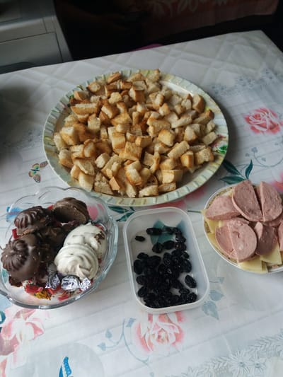 The image shows a table with a patterned tablecloth, potentially in a domestic setting in or near Бородулиха ауылдық округі, Kazakhstan, although this cannot be definitively confirmed from the image.
On the table are four containers of food:
1. A round plate filled with cubed bread.
2. A round bowl with sliced sausage and cheese.
3. A rectangular plastic container with dried black raisins.
4. A glass bowl with chocolate-covered candies and meringue.
There are no people visible in the frame, and no apparent actions are occurring.