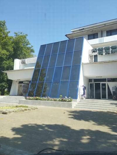 A bright, sunny day is depicted in this medium shot of a modern building with a large, angled glass facade. The glass structure, with its dark blue panes and blue framing, dominates the center of the image. In front of the building, there are several wide, light grey concrete stairs leading up to an entrance. A flower bed with pink and yellow flowers is situated in front of the stairs and along the base of the glass structure. To the left of the glass facade, trees with lush green foliage are visible. The building itself is white with flat rooflines. On the right side of the image, there's an entrance with glass doors, a sign that says "Coffee," and a vending machine. Above the entrance, on the second floor, several green tarps are draped over what appear to be outdoor chairs. There are no people actively engaged in any activity, but there are mannequins dressed in white and purple garments displayed on a rack near the entrance to the right. The scene appears to be a commercial or public building, possibly a hotel or a business establishment, in a location that suggests a warm, pleasant day.