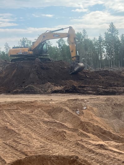 In Shapki, Russia, an excavator is operating on a construction site. The excavator, a SANY SY330H model, is positioned on a large mound of dark soil. Its arm is extended, and the bucket is digging into the soil. In the background, a dense forest of pine trees is visible under a partially cloudy sky. In the foreground, a section of the construction site is composed of lighter-colored sand with visible tire tracks from heavy machinery. There are no people visible in the image.