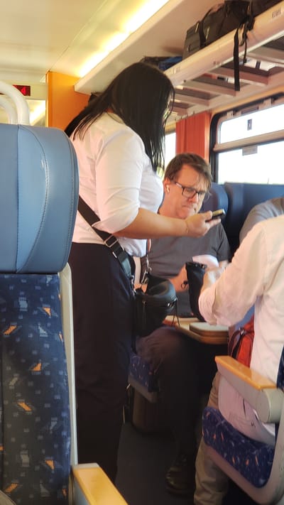 In an indoor setting resembling a train carriage, a woman with long black hair, wearing a white shirt and dark pants, stands with a black crossbody bag. A man in a grey t-shirt and glasses is seated, holding a phone. Another person, partially visible and wearing a white shirt, is seated to his right. The environment includes blue train seats with a patterned design and overhead luggage racks. The scene is likely within the city of Trenčín, Slovakia, based on the provided context.