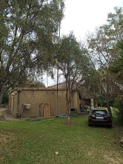 This media file depicts an outdoor scene in Midrand, South Africa, likely a residential or commercial property with a yard and a building. The primary subjects include a black Lexus SUV parked on a grassy area and a light brown, single-story building with a corrugated iron roof visible behind some trees. Several trees, some with dense foliage and others with more sparse branches, dominate the foreground and middle ground, suggesting a temperate or slightly humid climate.
The scene appears to be set during the daytime, with overcast or bright sky visible through the tree canopy. There are no people visible in the frame. The building has a textured, likely stucco or concrete, exterior. Various objects are placed around the building and yard, including a wooden shelf or panel mounted on the wall, a green trash bin, a mop and bucket, a coiled blue hose, and a long white pole with a green nozzle.
The license plate on the Lexus is visible and reads "ML 89 CX GP". The overall impression is one of a casual, slightly unkempt, outdoor environment, possibly a backyard or side area of a property. There are no obvious indications of a specific event or activity taking place, beyond the car being parked and some basic outdoor equipment being present. The weather seems to be mild, with no strong indicators of sun or rain.