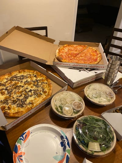 A bird's-eye view captures a wooden table laden with food from Andy's Pizza in Chevy Chase, Maryland. Two pizzas are visible: one with a classic red sauce and cheese, and another topped with mushrooms and cheese. Alongside the pizzas are two takeout containers of salad, one appearing to be a Caesar salad with parmesan cheese and croutons, and another a mixed green salad with feta cheese and what looks like beets, both served with dressings. A stack of stainless steel tumblers sits to the right of the pizzas, along with silverware and a receipt. In the foreground, a decorative paper plate with a floral design rests on the table. The background shows the interior of a dimly lit room, with glimpses of furniture, suggesting a residential setting. The overall scene depicts a casual meal, possibly a family dinner or gathering.