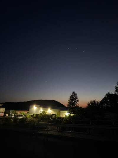 The image captures a twilight scene in Krumovgrad, Bulgaria, dominated by a gradient sky that transitions from a deep navy at the top to a soft orange and pink hue near the horizon. Several celestial bodies, likely planets or bright stars, are visible in the upper portion of the sky.

In the middle ground, a long, modern-looking building with bright lights illuminating its facade stands out against the darker landscape. It is surrounded by lush green foliage and trees. A white truck and a white van are parked in front of the building. A dark, silhouetted mountain range forms the backdrop to the building.

In the foreground, there appears to be a wooden structure or boardwalk with railings, suggesting an outdoor seating or viewing area. The overall scene conveys a sense of calm and transition from day to night.