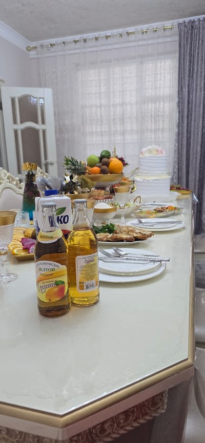 The image depicts a table set for a celebration, likely a meal or event, in what appears to be a residential dining room. The table is covered with a light-colored tablecloth with gold trim.

Prominently featured in the foreground are two glass bottles of beverages, one with a yellow label depicting a pear and the text "ЗЕЛТОН НАПИТКИ" (Zelton Drinks), and another bottle filled with a yellow liquid. A carton of what appears to be milk or juice is positioned between these bottles.

Various food items are visible on the table, including a tiered cake decorated with white frosting and pink flowers, a bowl of mixed fruits (pineapple, oranges, apples, kiwi), a plate of what looks like cooked meat or poultry, and a salad in a rectangular dish. Several plates, cutlery, and glasses are also arranged on the table, suggesting a formal setting. Decorative elements, such as gold-trimmed vases and a fruit bowl, add to the celebratory atmosphere.

In the background, a window is covered by sheer white curtains and heavier grey drapes. The room's interior, including furniture and wall trim, suggests a domestic environment.

While the image itself does not contain direct evidence of the city of Grozny, Russia, the provided prompt states this location. Therefore, inferring based on the prompt, this scene is set in Grozny, Russia. The overall impression is of a carefully prepared and decorated table, ready to host guests for a special occasion.