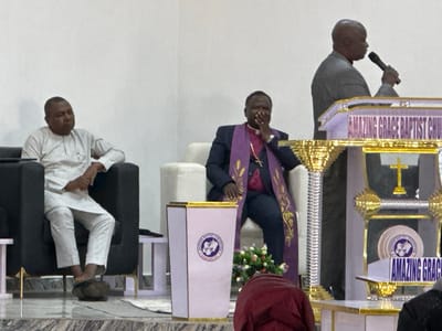 The image depicts an indoor religious gathering at Amazing Grace Baptist Church in Goni Gara, Nigeria. Three adult males are prominently featured, with a fourth individual partially visible in the foreground.
On the left, a man with dark skin and short dark hair is seated in a black armchair with gold accents. He wears a light-colored, long-sleeved tunic shirt with a small badge on the left chest, and matching trousers. His right foot, clad in a dark slipper, rests on a polished, marbled floor. He looks towards the right.
In the center, another man with dark skin and short dark hair, identified by his attire as a cleric or bishop, is seated in a light-colored armchair. He wears a dark suit jacket over a white clerical collar shirt, adorned with a purple stole embroidered with gold patterns. A gold crucifix necklace is visible. His right hand is raised to his mouth. He is also looking towards the right.
On the right, a third man with dark skin and a bald head stands behind an ornate, multi-tiered podium. He wears a grey suit jacket and holds a dark microphone to his mouth with his right hand, facing towards the left profile. The podium is light-colored (possibly purple or white) with gold trim and features "AMAZING GRACE BAPTIST CHURCH" inscribed on its upper front panel. A golden cross is visible on a lower shelf of the podium.
In the foreground, a tapered white pedestal with gold trim displays a circular logo of the "NIGERIAN BAPTIST CONVENTION," depicting a cross, an open Bible, and an atomic symbol, with the text "GO YE THEREFORE, AND TEACH ALL NATIONS." A small arrangement of multi-colored flowers is placed on the floor next to this pedestal. The upper back of a person clad in a dark red garment is visible in the lower center foreground.
The background consists of plain, light-colored walls. The flooring is polished and appears to be marbled or tiled. A small white rug or mat is visible near the feet of the man on the left.