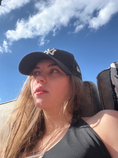 A young woman wearing a black New York Yankees baseball cap and a black tank top looks up and to the right, her face illuminated by sunlight. She has long, brown hair and is wearing a silver chain with a cross pendant. The background is a bright blue sky with scattered white clouds. She appears to be sitting outdoors, possibly on a couch or chair, as a sliver of a dark grey upholstered object is visible on the left and right sides of the frame.