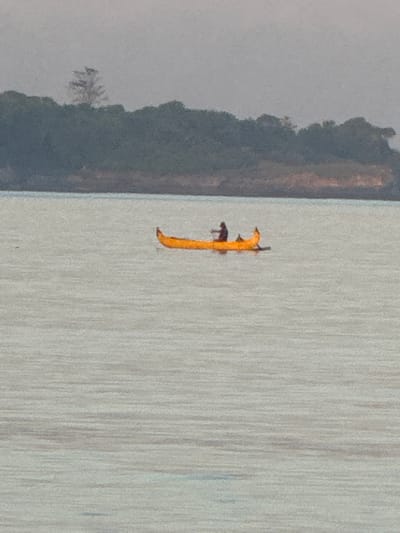 The image shows a portion of Jimbaran Bay in Indonesia, seen from an elevated position. The water is a light gray color with vertical streaking, possibly indicating surface texture or light reflection. A narrow, vertical yellow object, possibly a boat or surfboard, with some dark elements is positioned in the water. To the left is a strip of land with vegetation in shades of gray and brown. The overall image has a hazy quality.