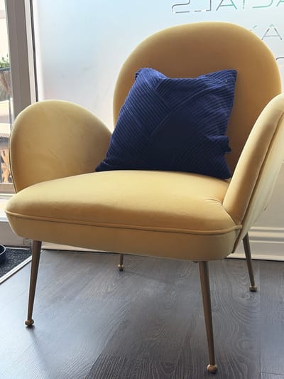 A close-up, eye-level shot shows a plush, mustard-yellow armchair with a dark blue, ribbed pillow resting on it. The chair has a rounded back and seat, with slender, gold-colored legs that end in small, spherical feet. The chair is placed on a dark gray, wood-grain laminate floor. In the background, a window with frosted glass reveals a blurred view of the exterior, with hints of greenery and sunlight. Above the window, a salon's branding is visible in reflective lettering, reading "2JAIDAS K". The overall impression is one of a well-appointed, modern interior. This scene is not in Torquay, United Kingdom.