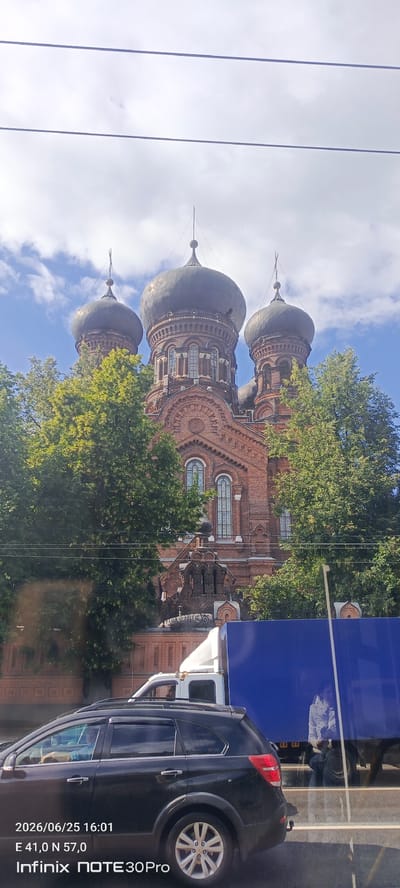 This media file captures an exterior view of a large brick building with multiple domes, suggestive of a church or cathedral. The building is constructed of red brick and features ornate architectural details, including arched windows and decorative brickwork. Several large, dark domes crown the structure, some topped with spires. Lush green trees partially obscure the lower and middle sections of the building, suggesting a daytime setting with ample sunlight.
In the foreground, two vehicles are visible. A dark-colored SUV is parked in the lower left, and a white truck is positioned behind it, partially obscured by the SUV. A large blue truck, possibly a semi-trailer, is parked to the right, its side reflecting the surroundings. A person, dressed in a light-colored jacket, is seated inside the blue truck. The scene appears to be on a street, with visible road markings and possibly a traffic lane.
The image is dated "2026/06/25 16:01" and provides GPS coordinates "E 41,0 N 57,0". The text "Infinix NOTE 30 Pro" is also present, indicating the device used to capture the image. The sky above is a mix of blue sky and white clouds, consistent with a partly cloudy day. The overall impression is a snapshot of a moment in an urban or semi-urban environment, focusing on the architectural landmark and the surrounding vehicles. The presence of traffic and the daytime lighting suggest a normal, active day.