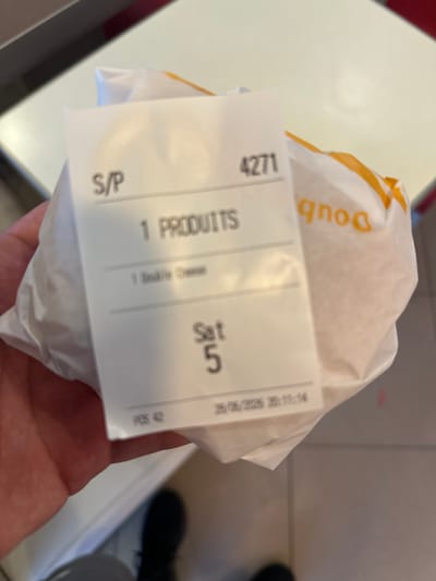 A person is holding a food item wrapped in white paper with a yellow and orange logo visible on the paper. Attached to the food item is a white receipt or sticker. The receipt is printed with black text and has several sections. The top left corner shows the number "4271". Below that, it reads "S/P". To the right of "S/P", it says "1 PRODUITS". Further to the right, the day is indicated as "Sat 5". A date and time stamp is visible in the top right, reading "26/06/2026 20:11:14". Below the date and time, it shows "POS 42". There is also some faded text that is difficult to read. The food item appears to be a burger or sandwich. The background is blurred, but suggests an indoor setting, possibly a fast-food establishment or a casual dining environment. The lighting appears to be artificial.