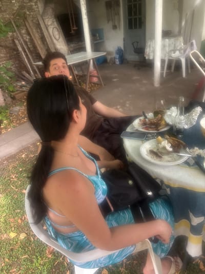 An overhead shot reveals a woman in a light blue patterned swimsuit sitting at a table. Nearby, a man in a brown shirt looks up at the camera. The table is cluttered with plates of food, utensils, and a bottle of Corona beer. A black bag sits on the table. The background shows a weathered exterior with a porch, chairs, and some greenery. The photo is taken outdoors on a sunny day.