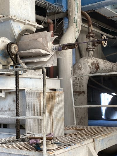 The image depicts an industrial setting with heavy machinery and piping, likely within a manufacturing or processing plant. The machinery is predominantly made of metal, with some parts covered in dust or residue, giving them a worn and weathered appearance. Large pipes, some with a rusty exterior, snake through the scene, connecting various parts of the equipment. The overall environment appears to be gritty and industrial, with a focus on functional, heavy-duty equipment. There are no people visible in the image, and the scene appears static, suggesting a moment captured during a period of operation or maintenance. The location is specified as Police, Poland, indicating an industrial zone within that city.