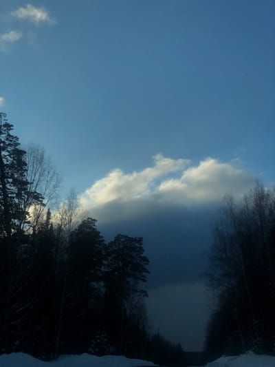 The image captures a dramatic winter landscape, likely in Izvestkovyy, Russia, with a striking contrast in the sky. The left side of the frame features a clear, bright blue sky dotted with scattered, sunlit white clouds. This transitions abruptly to the right, where a massive, dark, and dense cloud formation dominates the sky, casting deep shadows below.

A dense line of trees, including both evergreen and bare deciduous varieties, forms a dark silhouette along the horizon. These trees are heavily shadowed, particularly on the right side where they are under the dark cloud, with hints of snow visible on some branches and the snowy ground. On the far right, a snow-covered path or ground is partially visible through the trees.

The scene depicts a clear, cold winter day with a notable weather event or a rapidly approaching weather front. The time of day appears to be daytime, possibly late afternoon, given the bright areas and deep shadows. No people, discernible activities, or visible text are present in the image. The powerful contrast between the bright sky and the ominous dark clouds, along with the snow-covered forest, evokes a sense of awe and natural drama.