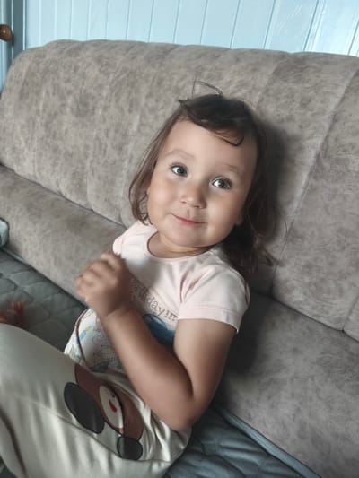 A young child with light brown hair and green eyes is sitting on a beige couch. The child is wearing a light pink t-shirt with a graphic design and light-colored pants with a dog-like design. The child is looking towards the camera with a slight smile. The background shows a light blue wall with horizontal panels. The setting is indoors, likely a home, and the image does not provide any specific information to determine the location within the city of Burayevo, Russia.