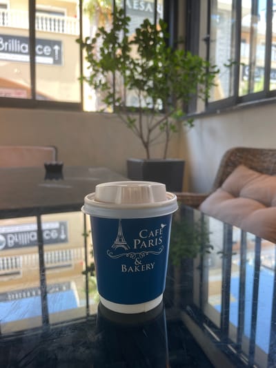 A medium shot captures a disposable coffee cup with a blue exterior and a white lid. The cup features an illustration of the Eiffel Tower and the text "Cafe Paris & Bakery" in white lettering. It sits on a dark, reflective surface, likely a table, in what appears to be an outdoor seating area.

In the background, a potted plant with green leaves is visible, suggesting an interior or covered outdoor space. Behind the plant, a window reveals a glimpse of a building facade with balconies and signage. One sign is partially visible and reads "Brillia ce 1", indicating a commercial establishment, possibly a shop or restaurant. The overall lighting suggests a bright, daytime scene. The location is identified as Trikomo, Northern Cyprus.