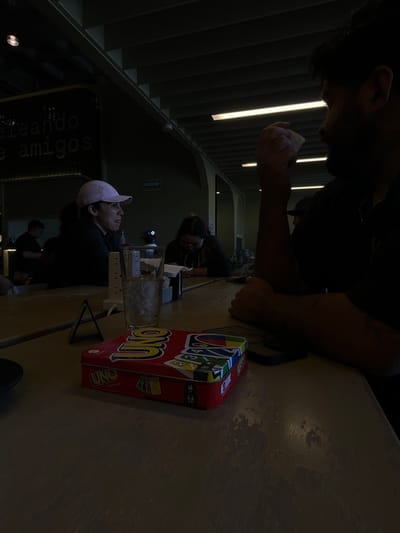 A group of people are gathered around a table. One man is seen from above with his hands clasped together near his face. At the table, there is a box of UNO cards, a glass of water, and electronic devices. Other individuals sit around the table facing in the same direction, and one man is wearing a pink cap. The background features a building with a lit staircase.