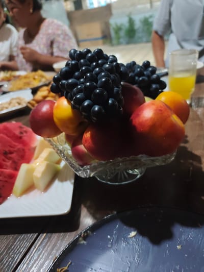 A table is set for a meal, with a fruit bowl as the centerpiece. The bowl, made of clear, patterned glass, holds a vibrant assortment of fruits, including dark purple grapes, red and yellow nectarines or peaches, and perhaps an orange. To the left of the fruit bowl, a white plate displays sliced watermelon and chunks of melon, possibly cantaloupe or honeydew.

In the background, two people are visible, seated at the table. One person, on the left, is wearing a patterned top and has short, dark hair. The other person, on the right, is wearing a light-colored shirt. The setting appears to be indoors or under a covered area, suggested by the presence of furniture and a glimpse of greenery outside. A glass of yellow liquid, possibly juice, sits on the table near the fruit bowl. The overall scene suggests a gathering or a meal, possibly a family get-together. The lighting indicates it might be daytime or early evening. The image lacks any visible text.