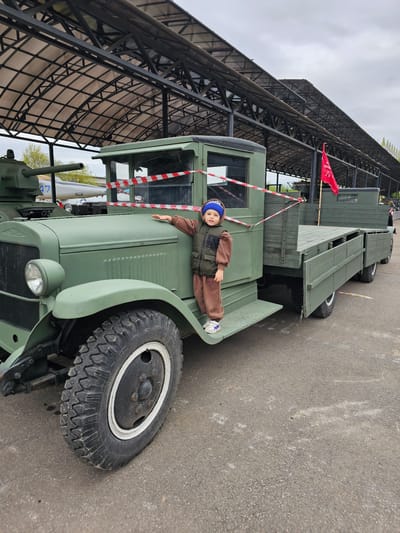 A young child wearing a blue hat, brown sweat suit, and a dark vest is lying on the fender of a vintage green military vehicle. The vehicle appears to be a truck and has a red flag with white writing attached to the rear. The vehicle is parked beneath a large metal structure, and there is a white and red striped barrier visible near the cab of the truck. The location is likely Arkhangel’skoye, Russia. The ground surface appears to be asphalt.