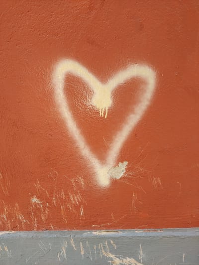 The image shows a close-up, full-frame shot of a red wall with a white spray-painted heart on it. The heart is imperfect, with drips of white paint running down from the top curve. The wall itself is textured and appears weathered, with visible scratches and scuffs, particularly on the lower portion. A grey concrete base runs along the bottom of the wall, also showing signs of wear and tear. The lighting is even, suggesting an overcast day or diffused indoor lighting. There are no people or other identifiable objects in the frame. The image is taken in Perpignan, France.