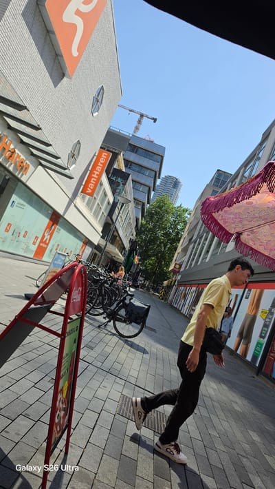 A man in a yellow polo shirt and black pants walks down a sun-drenched city street in Rotterdam, Netherlands. He wears white sneakers with brown accents and carries a black bag over his shoulder. His gaze is directed upwards and to his left, seemingly lost in thought.
The scene is bustling with activity. In the foreground, a rack of bicycles is parked along the sidewalk. A red A-frame sign, possibly for a restaurant or promotion, stands near the bikes, displaying an image of pizza. Beyond the man and the bikes, the street is lined with modern buildings, some featuring large windows. A construction crane is visible in the distance, hinting at ongoing development in the city.
The sky is a clear, bright blue, indicating a sunny day, likely during midday due to the strong shadows cast on the pavement. The pavement itself is composed of grey rectangular tiles, and a grate is visible on the sidewalk. Above the man, a colorful, fringed fabric hangs suspended, adding a touch of vibrant artistry to the urban landscape.
The overall impression is one of a typical, lively urban day in Rotterdam, with a mix of commerce, transportation, and everyday life unfolding under a pleasant sky. The man's posture and expression are neutral, suggesting a moment of personal reflection amidst the public space. Visible text includes "VanHaren" on an orange sign, and "Galaxy S26 Ultra" watermarked in the bottom right corner.