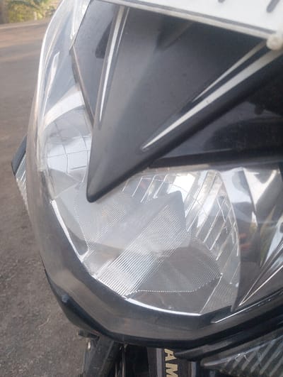 A close-up, angled view depicts the front section of a black motorcycle, presumably located in Cibeureum, Indonesia. The primary subject is the motorcycle's headlight, encased in a clear plastic lens with internal textured reflector patterns consisting of horizontal and diagonal ridges. Surrounding the headlight is the black plastic fairing, featuring an angular design and incorporating white or light-colored linear accents. A partial view of a registration plate mount is visible at the top edge of the image. Below the headlight, a vertical black surface displays partial lettering, "YAMA," suggesting the brand of the vehicle. The background, out of focus, shows a light-colored paved road surface, indicating the motorcycle is stationary on a street or similar thoroughfare under natural daylight conditions. No individuals, specific actions, or other definitive environmental structures are discernible.