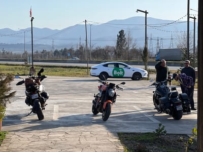 The image depicts an outdoor scene in Argveta, Georgia, under a clear daylight sky with distant mountains. Three motorcycles are parked on a grey asphalt surface with white markings. From left to right: a silver/grey adventure motorcycle (GEORGIA F 0780) equipped with a brown luggage bag, an orange naked sport bike, and a black cruiser motorcycle (GEORGIA BA 1494) fitted with saddlebags. Three adult males are gathered around the black cruiser motorcycle. One male, wearing a plaid shirt, is bent over the handlebars of the black motorcycle, seemingly inspecting it. A second male, in a dark green long-sleeved shirt, stands with his left hand on his head. A third male, wearing sunglasses and a dark long-sleeved shirt, stands upright, observing the interaction. In the mid-ground, a white sedan with a prominent green "Bolt" logo is parked. Behind the parking area is a multi-lane highway where a light-colored sedan and a blue truck with a grey trailer are in motion. A black pole bearing the Georgian flag is visible on the far left. The background features utility poles, power lines, bare trees, and a range of mountains. The immediate foreground shows a portion of a stone paver patio.