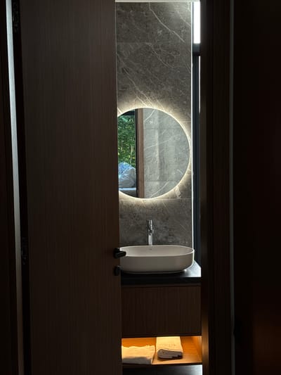 The image depicts a modern bathroom with a minimalist design, located in Bakovići, Montenegro. A dark wooden door is partially open, revealing a glimpse into the room. Inside, a sleek white countertop with a vessel sink is visible, complemented by a chrome faucet. Above the sink, a circular mirror with integrated LED lighting creates a halo effect. The wall behind the sink is clad in large-format grey marble tiles, adding a touch of luxury. A tall, thin window or light fixture runs vertically along the right edge of the tiled wall, allowing a sliver of natural light to enter. Below the countertop, a wooden cabinet houses neatly folded white towels illuminated by a soft, warm light from an internal shelf. In the reflection of the mirror, greenery and what appears to be a light-colored comforter or draped fabric can be seen, suggesting an outdoor view or an adjacent living space. The overall ambiance is one of sophisticated tranquility.