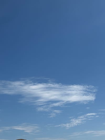 A vertical, medium-shot photo shows a bright blue sky with a long, thin, wispy cloud stretching horizontally across the middle of the frame. The cloud appears to be composed of cirrus formations, indicating fair weather. The bottom edge of the image contains a small, dark, textured area that appears to be the top of a building or structure, suggesting it is a terrestrial scene. The location is Ostrówek, Poland.