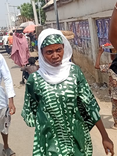 This image depicts a bustling street scene in Ogaminan, Nigeria. In the foreground, a dark-skinned woman with a direct gaze is positioned slightly off-center. She wears a loose-fitting green garment with light green patterned designs, a light green patterned headscarf, and an overlying white headscarf. Her left arm is visible, hanging by her side.
To the immediate left of the central figure, the lower torso and legs of an individual in a light blue long-sleeved shirt, light-colored shorts, and grey flip-flops are partially visible. Further back and to the left, a woman in a flowing pink or light purple robe is observed walking away from the viewer, appearing to carry an item. Behind her, a small, bare-chested individual with dark, curly hair is positioned closer to the ground. Additional indistinct figures and a vehicle are visible further down the paved street, suggesting ongoing movement and traffic.
On the right side of the frame, a light-colored concrete wall is visible, covered with multiple political campaign posters featuring male figures in suits and partially visible text, including "PERCEN". Several large, light-brown sacks are stacked against this wall. The arm and shoulder of a person are partially visible on the far right, holding a tray with sliced red produce (consistent with tomatoes) balanced on their head, indicating a street vendor. A dark, vertical street pole stands near the wall. The scene is illuminated by bright daylight.