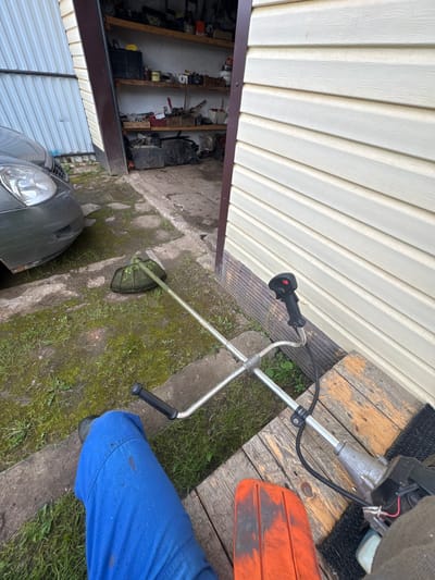A first-person perspective shot shows a person wearing blue work pants, sitting on a wooden platform outside a garage or shed. In the foreground, to the left, is the person's leg and part of a grass trimmer, partially obscured by the person. The grass trimmer is silver and black, with a black handle and a red button. Further back, to the left of the trimmer, is the front of a dark gray car. The garage or shed has a light-colored, horizontal siding and an open doorway revealing shelves stocked with various items, including containers, tools, and bags. The ground is a mix of concrete, dirt, and patchy green grass, with some puddles indicating recent moisture. The overall scene suggests a residential or utility area, possibly in Borodulino, Russia, where yard work is either about to commence or has recently been completed.