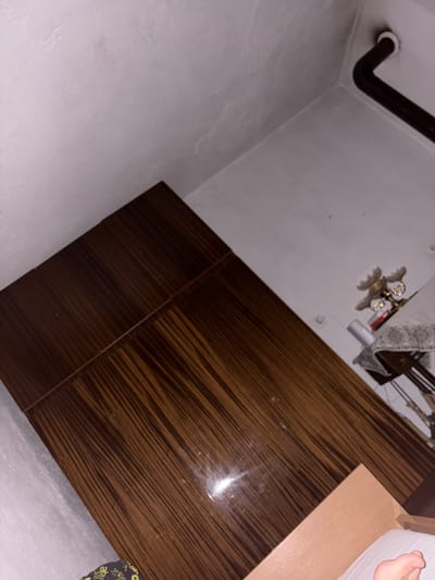 This image shows a close-up view of a wooden surface, possibly a table or a floor, with a distinctive striped grain pattern in shades of brown. The surface appears to be indoors, with a white wall visible in the upper left corner and a dark pipe running along the top edge of the frame. In the background, a small object that resembles a decorative lamp or chandelier can be seen on a surface covered with a patterned cloth. The overall lighting is dim, suggesting an indoor environment at night or with limited light. There is no clear indication of the specific location within the city of Kran, Bulgaria.