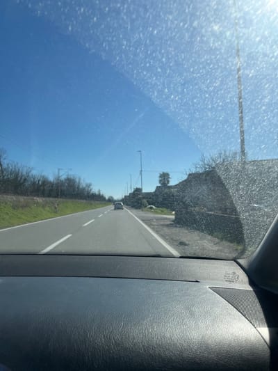 The image depicts a daytime scene from the interior of a vehicle, looking forward through a windshield. The windshield exhibits numerous dirt specks and streaks, particularly concentrated in the upper right quadrant, with sunlight causing significant glare.
The immediate foreground shows a dark, textured dashboard. In the distance, a paved two-lane road extends into the frame. The road features a solid white line on its right edge and a dashed white line separating the two directions of travel. A light-colored, possibly white or silver, small car is visible in the far lane, moving away from the viewer.
To the left of the road, a green grassy embankment is present, bordered by a dense line of bare trees or bushes. Overhead, utility wires or power lines are visible running parallel to the road. A small, reddish-orange marker pole is situated on the embankment.
To the right of the road, a rough, unpaved shoulder transitions into an area with what appears to be a low, dark-colored stone or concrete wall or structure, partially obscured by leafless vegetation. Several tall streetlights with black poles are spaced along this side of the road, receding into the distance. The sky is clear and bright blue, indicating sunny weather. No people are discernible in the scene. The location is Bistrica, Montenegro.