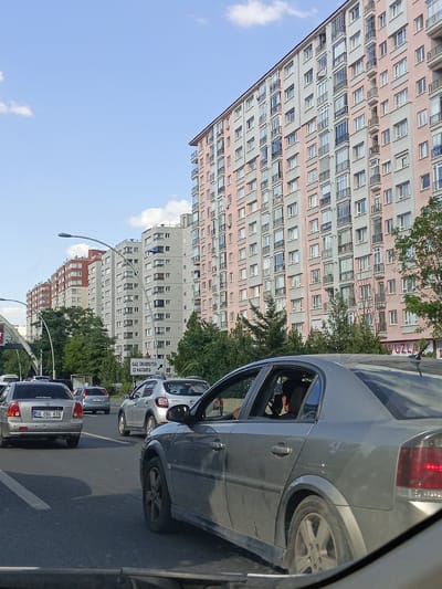 This media file depicts a street scene in Yenimahalle, Turkey, likely during daytime given the clear sky and shadows. The weather appears to be fair. The foreground is dominated by the rear and side of a silver sedan, partially obscuring the view of other vehicles in traffic. The car has a license plate that reads "06 EBG 939".

Behind the vehicles, a row of apartment buildings stretches along the street. The most prominent building on the right is tall and has a pink and grey facade, with numerous balconies and windows. To the left, there are other apartment buildings, some with light grey exteriors and others with red and white accents. Trees and greenery are visible between the buildings and along the roadside, suggesting a somewhat urban but also green environment.

Traffic appears to be moving, though possibly congested. Several cars are visible in different lanes. A street sign is positioned on the right side of the road, which reads "GAZİ ÜNİVERSİTESİ [symbol of hospital] HASTANESİ 130 m." This indicates that Gazi University Hospital is nearby. The overall atmosphere seems to be a typical urban commute. The time of day is likely afternoon or late afternoon due to the angle of the sun and the brightness of the sky.