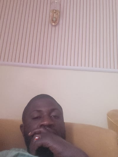 A male individual is depicted in an indoor setting in Babawa, Nigeria, appearing to be a selfie. The subject, a dark-skinned male with short hair and a beard, is positioned centrally, viewed from the chest up. His right hand, adorned with a ring featuring a reddish-orange stone on the ring finger, is partially covering his mouth and chin. He is wearing a light green or teal shirt and is seated on a light brown or tan upholstered chair or sofa, partially visible to his right and beneath him. The background consists of a plain, light beige wall. Above the wall, a ceiling section is visible, composed of light-colored, vertically ridged panels, separated from the wall by a white trim. A clear glass, wall-mounted light fixture with a gold-colored base is affixed to the ceiling panels in the upper center.