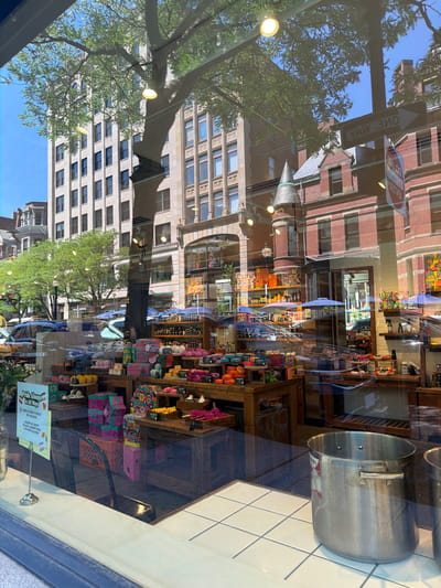 This is a photograph of a shop window display reflecting an urban street scene. The reflection shows buildings with red brick and stone facades, trees with green foliage, and a clear blue sky. A "ONE WAY" street sign is prominently reflected, with the text "YAW 3MO" visible due to the reflection. The shop interior, also visible, displays shelves stocked with various colorful items, possibly bath products or gifts, including soaps, boxes, and accessories. A large silver pot is also visible inside the shop. The overall impression is a sunny day in a city, with the reflections creating a layered visual experience of the exterior and interior. The text visible includes "ONE WAY," "YAW 3MO," and a sign in the shop window that reads "Pride Always in All Ways" with additional smaller text below. The lighting suggests it is daytime, likely late morning or afternoon.